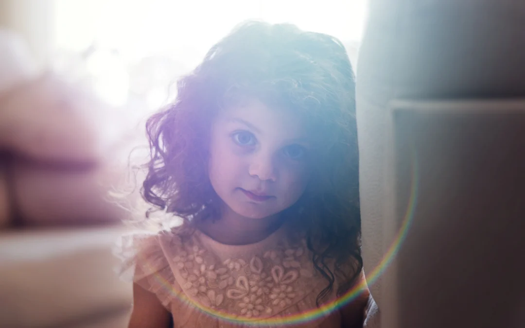 A young girl with dark curly hair looks directly at the camera with a soft expression. A sunbeam is coming from behind her and leaves a circular impression on the camera